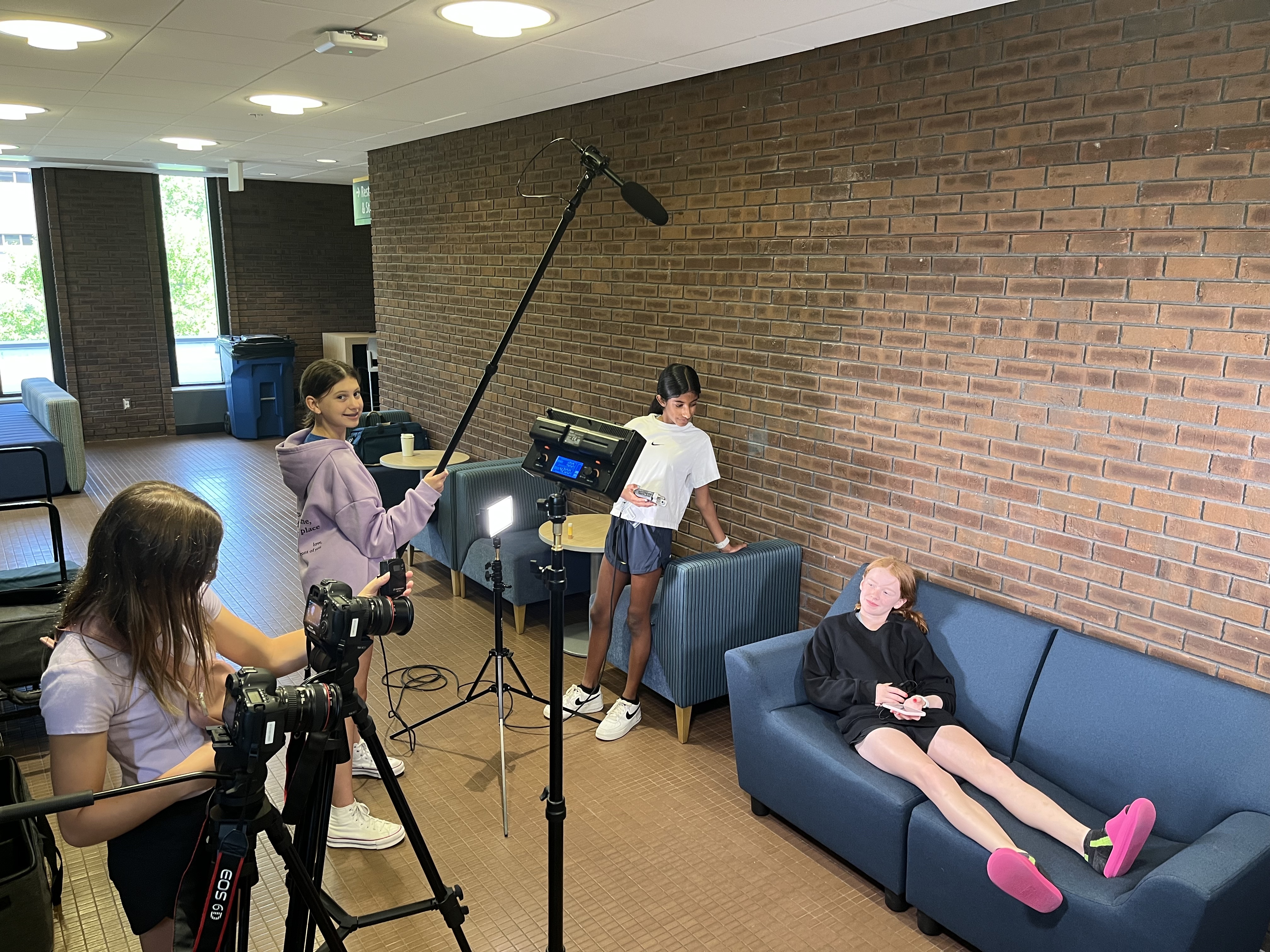 Four young students on a production set: One girl is behind a camera, one girl is holding a boom pole, one is checking audio levels and the fourth is on camera