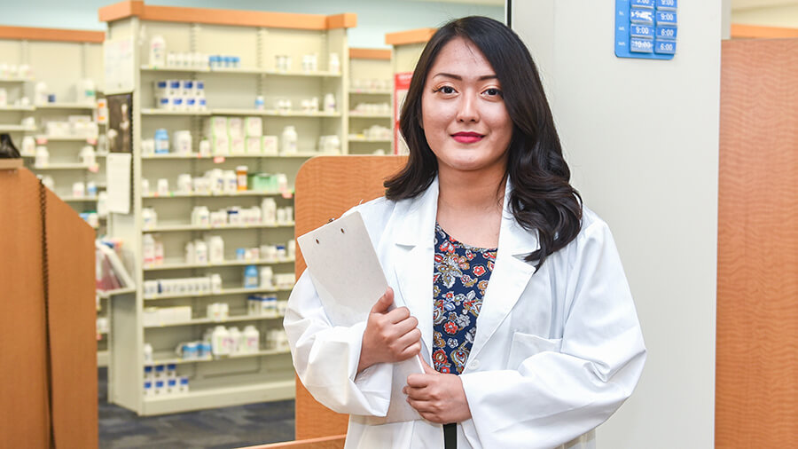 A woman wearing a lab coat and holding a clipboard smiles.