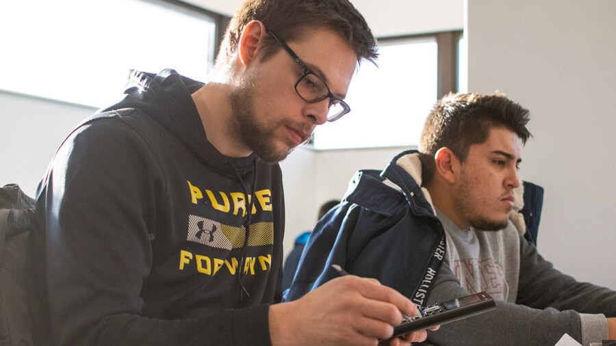 A student uses a calculator next to a student working on a computer.