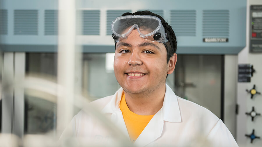 A student wearing a lab coat and safety goggles smiles.