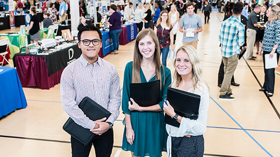 Three students pose for a photo holding portfolios during a career fair.