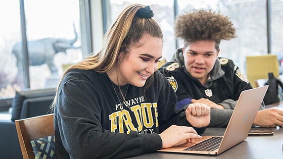 A student registers for classes on her laptop while another looks on.