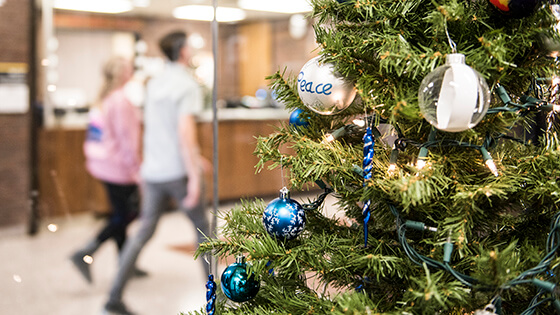 A student holds a gift while standing in front of a Christmas tree.
