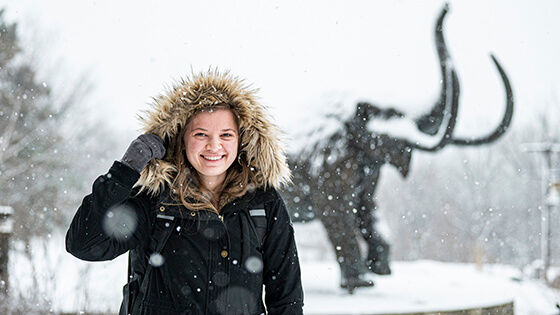 A student walks to class in the snow with the mastodon statue in the background.