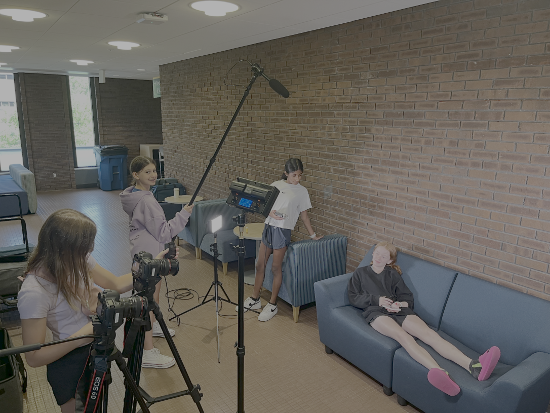 Four Students hold camera, light and audio equipment as they film a video in a library hallway.