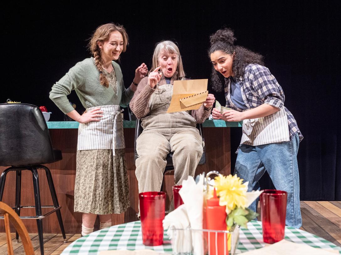 Three actresses on stage in a diner set are looking at a paper together. They look happy and surprised.