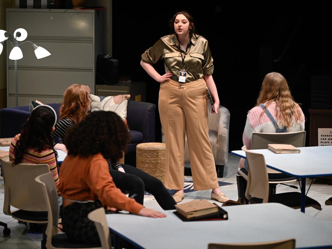 Actors in a play in a high school set, Four students are facing away from the camera towards a woman speaking to them.