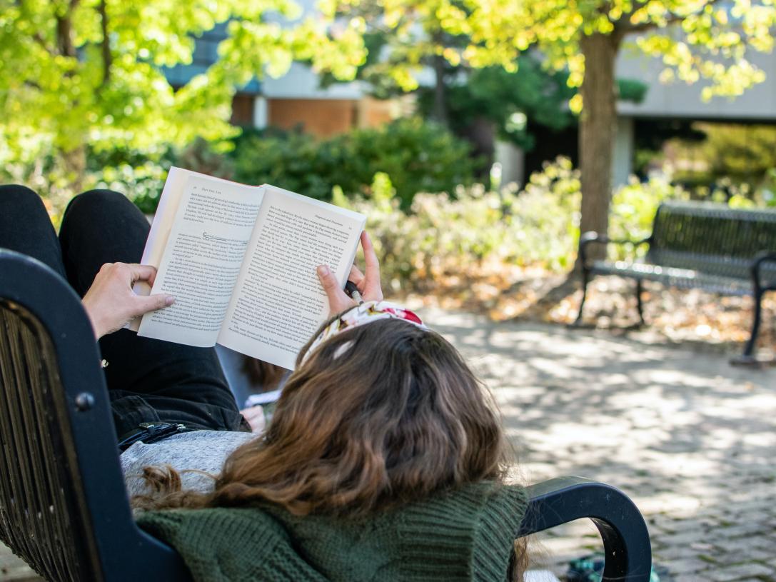 Student laying on a bench reading a book.