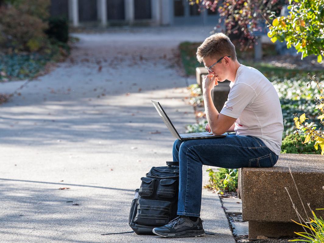 Student works on a laptop on a bench outside on campus.