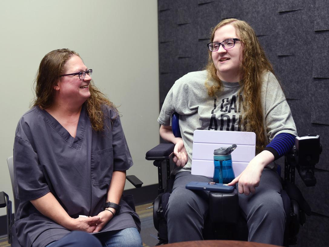 Elizabeth Farlow is seated in a wheelchair and speaking to a friend.