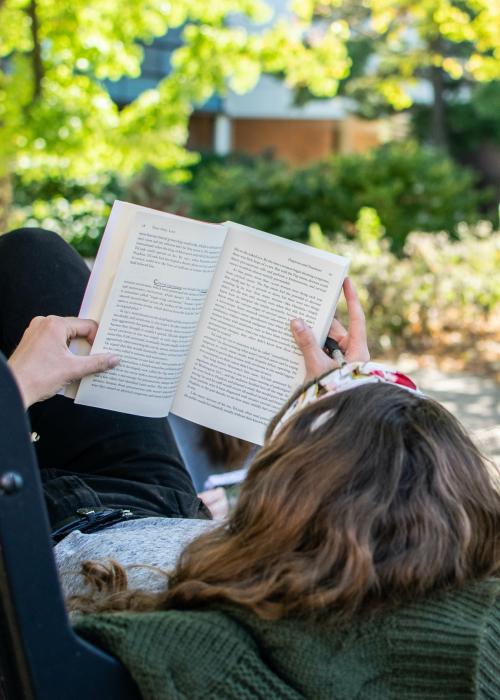 Student laying on a bench reading a book.