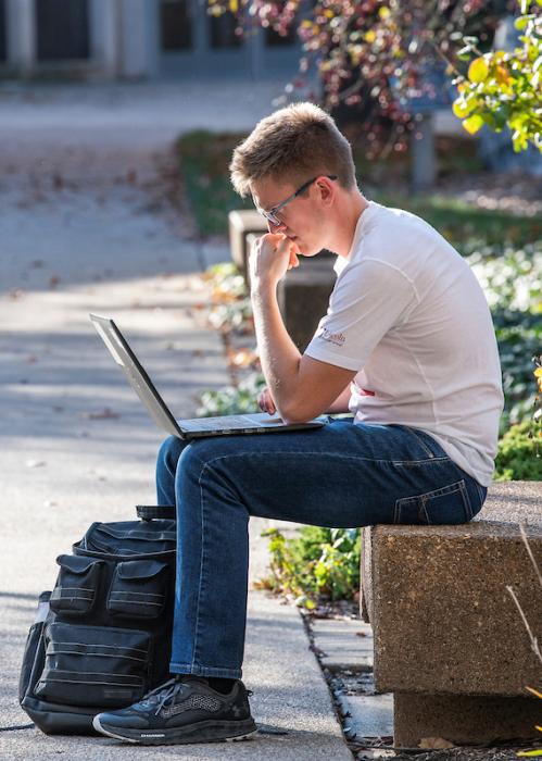 Student works on a laptop on a bench outside on campus.