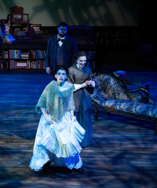 Actors on stage in dramatic blue lighting a woman looks scared, another woman is behind her looking mischevious, there is a man in the background observing, and a woman to the side in a rocking chair