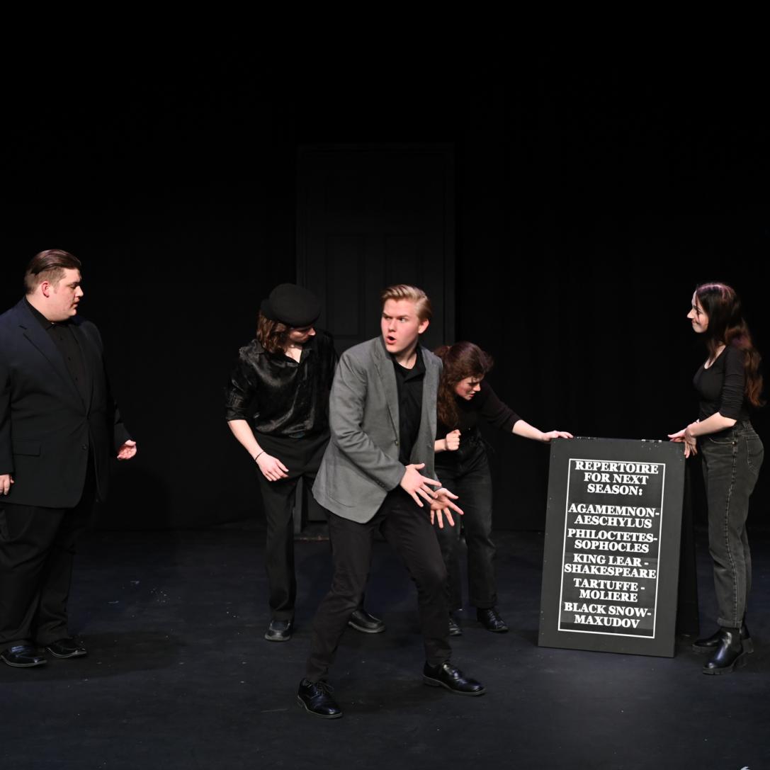 Actors in a black box theatre scene, several are setting up a sign in the background while a man gestures towards it with an exagerated expression of concern