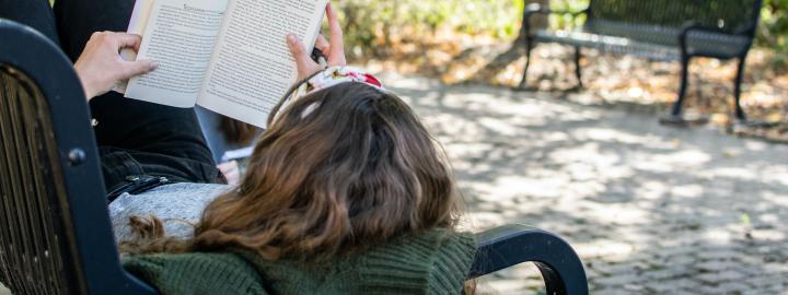 Student laying on a bench reading a book.