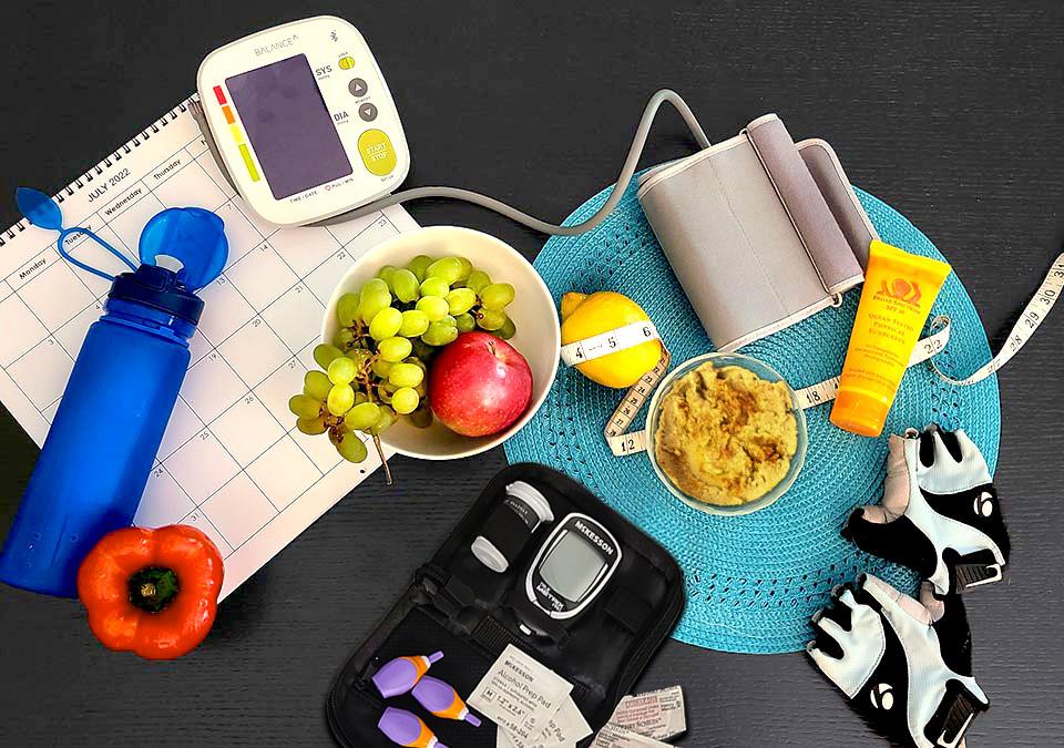 Healthy items to help monitor diabetes: glucose monitor, blood pressure monitor, fruits, an orange bell pepper, bowl of hummus, reusable water bottle, tape measure, and weight lifting gloves on top of a calendar and blue placemat on a gray table.
