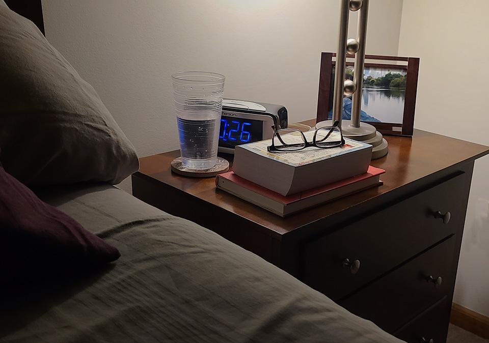 Photo of bed and softly lit lamp on night stand with books, reading glasses, clock, glass of water and picture in a frame