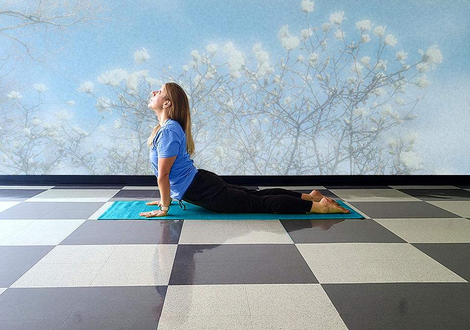 Female yoga instructor doing the cobra yoga pose on a turquoise mat in front of a wall with a muted mural of whitle magnolias and a blue sky.