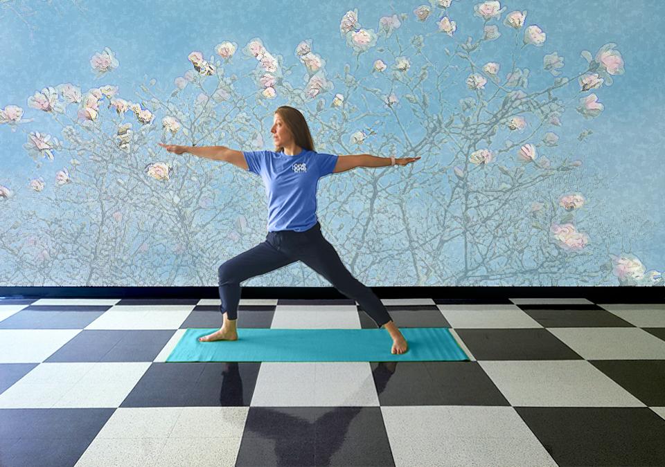 Female yoga instructor in a periwinkle t-shirt and black pants showing the warrior pose with a muted light blue wall with magnolias.