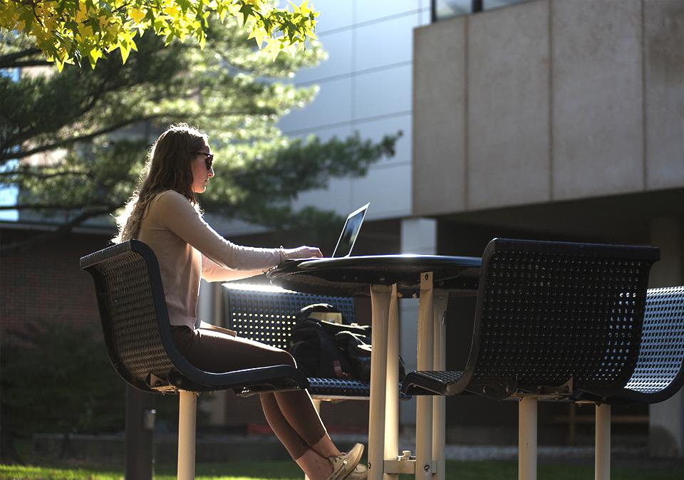 Student sitting outside at a bistro table while working on computer.