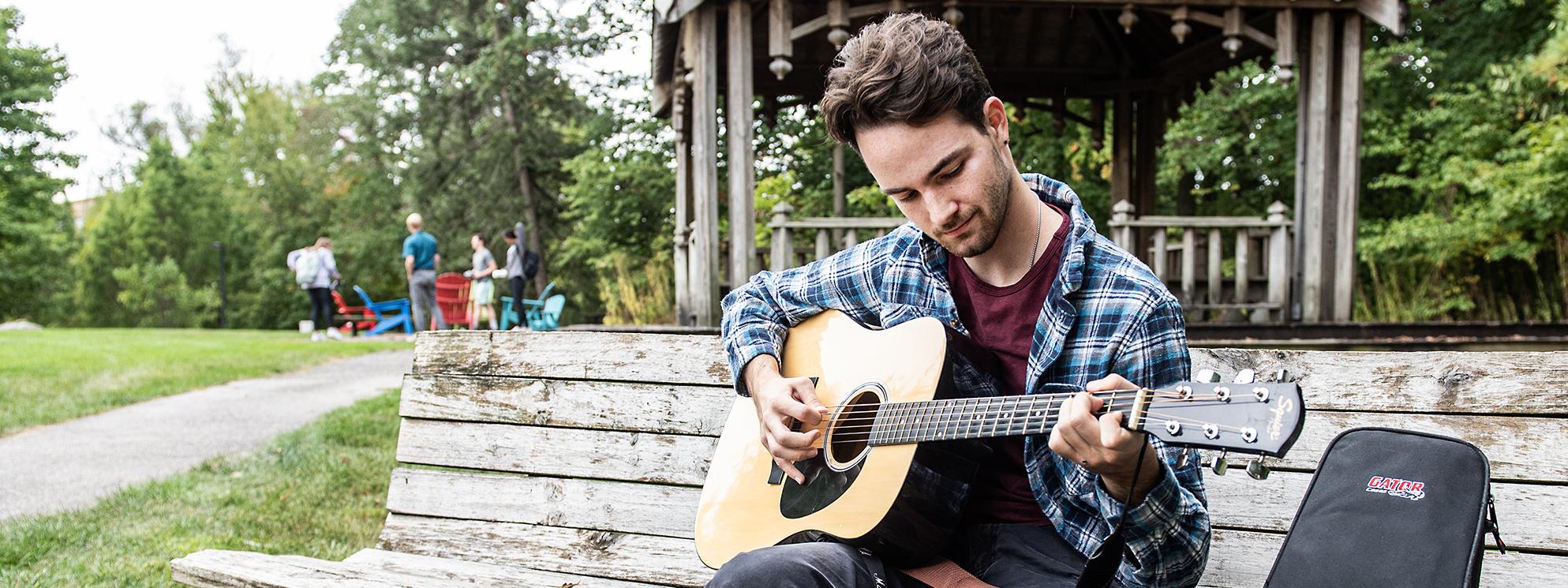 Student plays a guitar outside on campus while sitting on a bench.