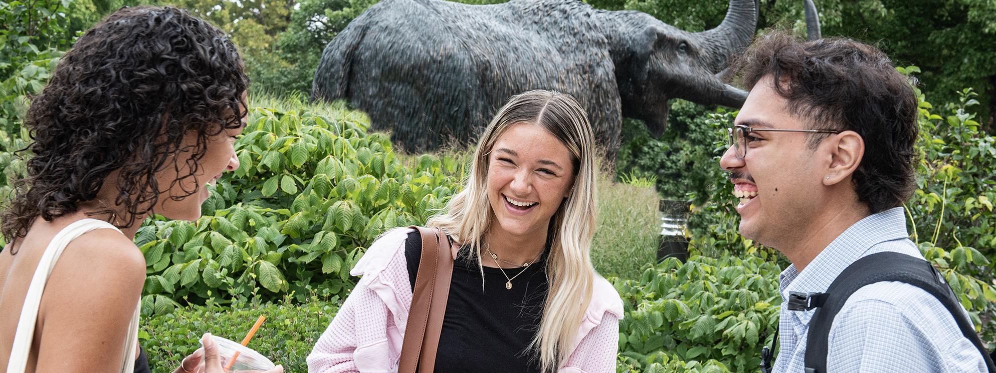 Three students laugh and chat outside in front of the Mastodon statue.