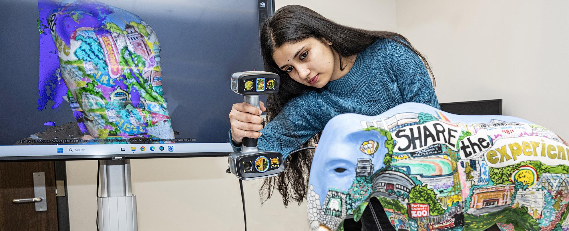 A student uses a handheld scanner to digital copy the design of a painted Mastodon statue.