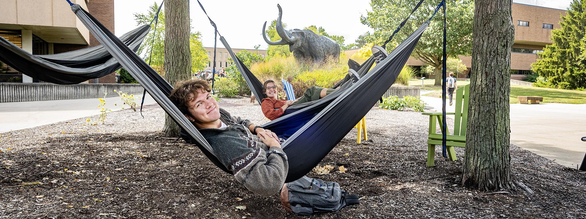 Students lounge in hammocks on campus in front of the Mastodon statue.