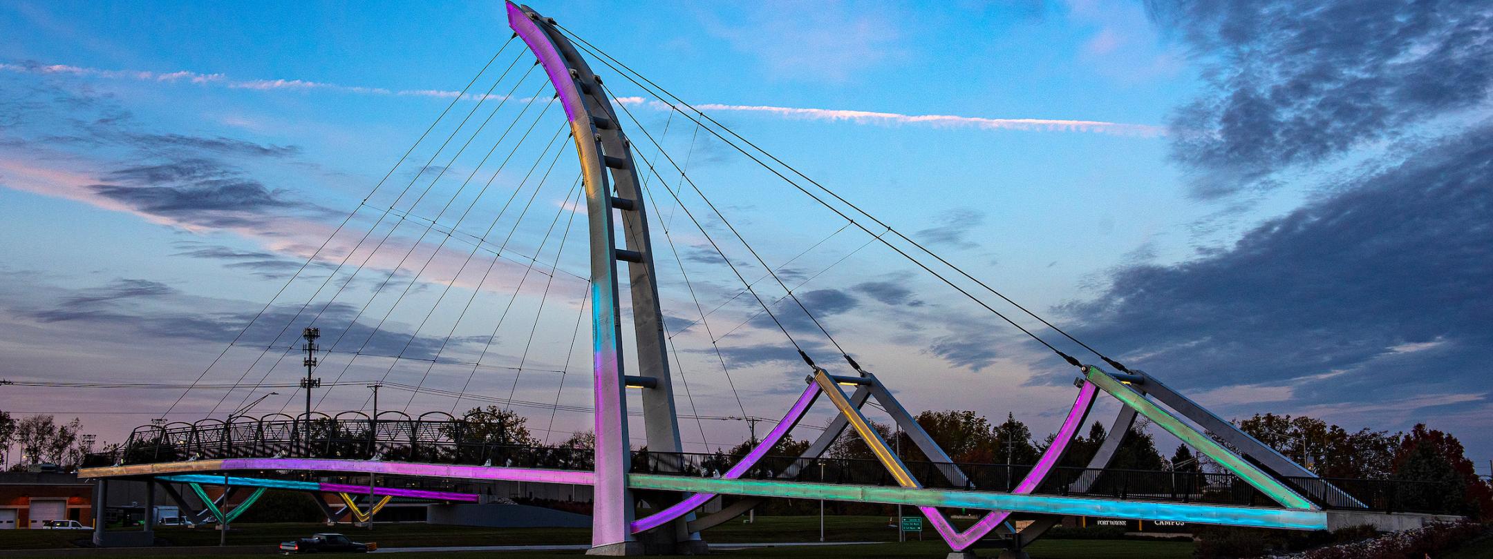 Image of PFW foot bridge with multi-colored lights at dusk.