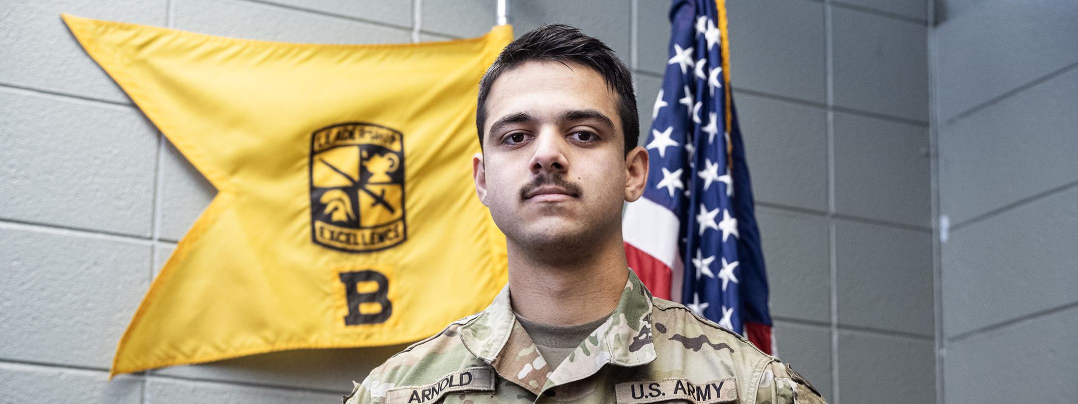 Tanis Arnold is wearing an Army uniform and standing in front of two flags