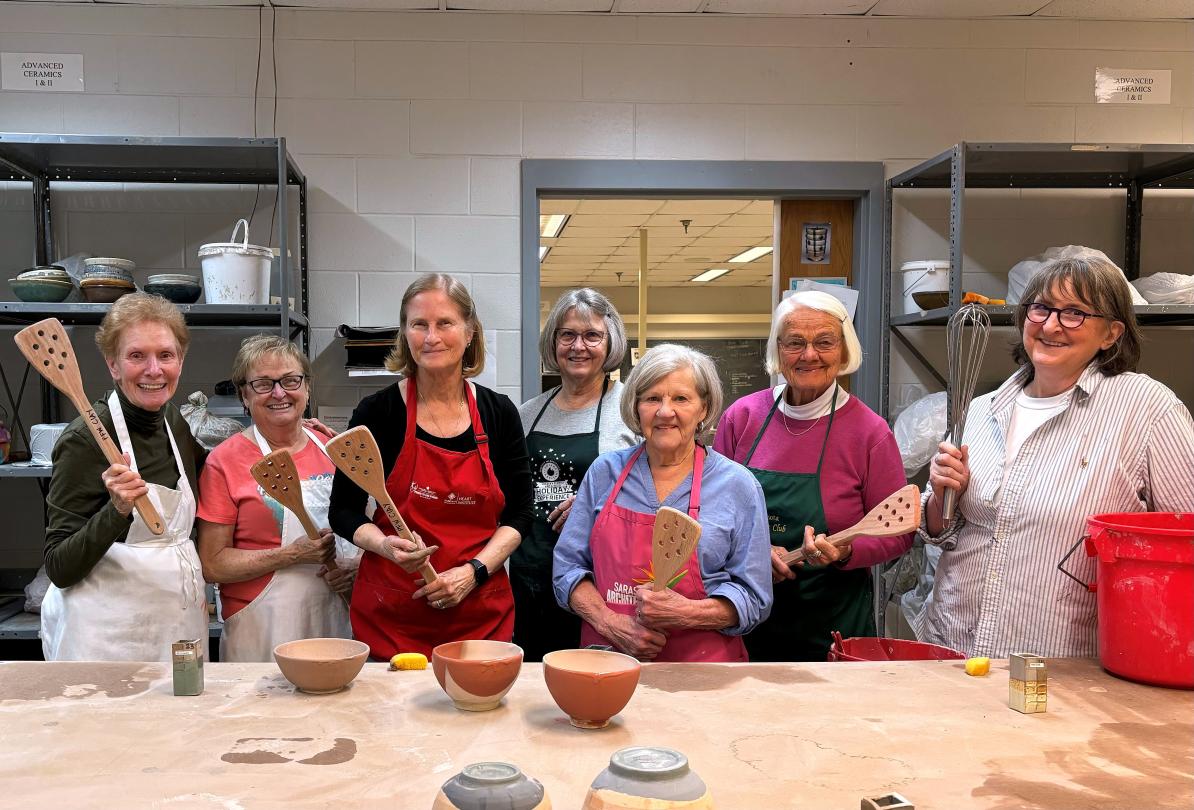 8 smiling ladies holding wooden tools during a pottery event. FRIENDS of the University