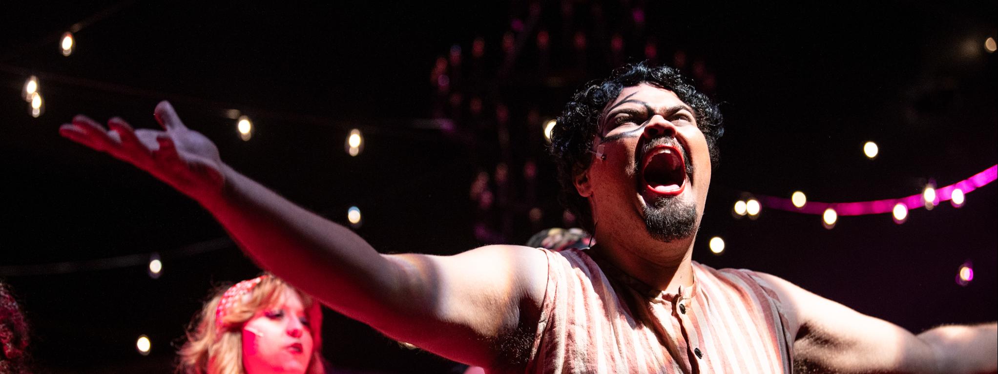 Emcee singing with his arms up from Cabaret, a dancer is visible behind him in dramatic red lighting