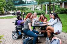 A group of laughing students sitting at a table outside the student union. One of the students is in a wheelchair.