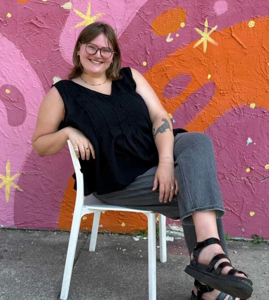 gwenna photoshoot, sitting in chair, behind a pink and orange outdoor mural