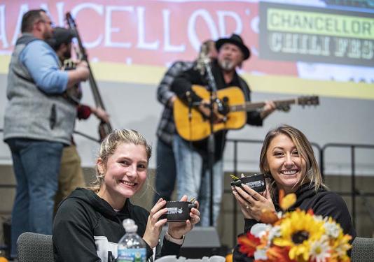 Two women are holding up mugs of chili. A bluegrass band is playing in the background.
