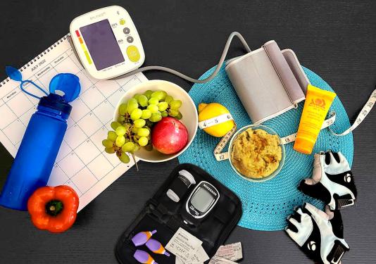 Healthy items to help monitor diabetes: glucose monitor, blood pressure monitor, fruits, an orange bell pepper, bowl of hummus, reusable water bottle, tape measure, and weight lifting gloves on top of a calendar and blue placemat on a gray table.