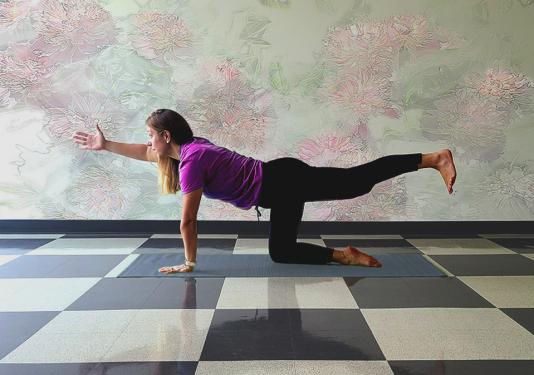 Health coach demonstating the bird dog yoga pose on a gray mat with pastel muted wall in the background and a white and black checkered floor