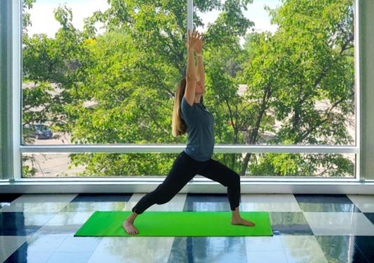 Health coach demonstrating warrior pose in front of windows with trees in the background