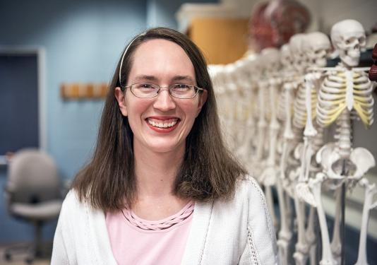 Regina Trout is standing in front of a row of human skeletons in a biology lab