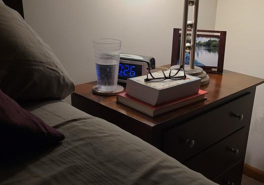 Photo of bed and softly lit lamp on night stand with books, reading glasses, clock, glass of water and picture in a frame