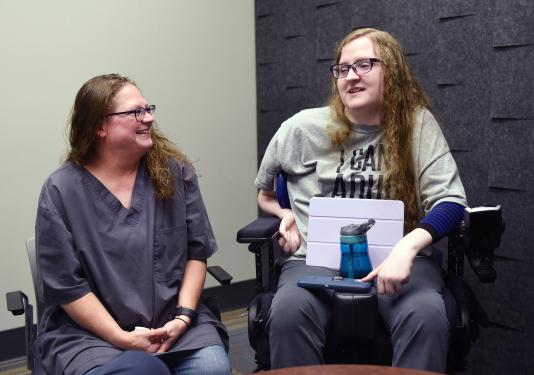 Elizabeth Farlow is seated in a wheelchair and speaking to a friend.