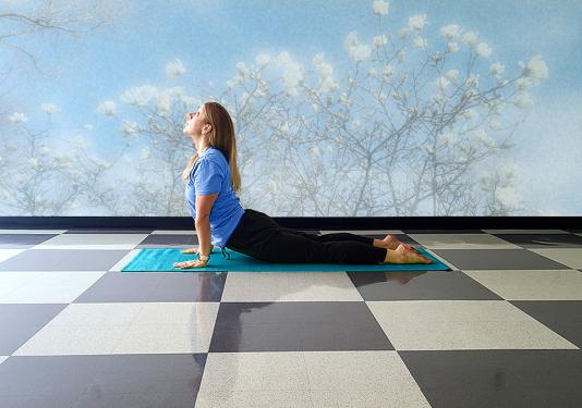 Female yoga instructor doing the cobra yoga pose on a turquoise mat in front of a wall with a muted mural of whitle magnolias and a blue sky.