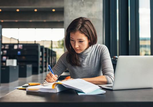 Shows a person in a library with labtop and books studying.