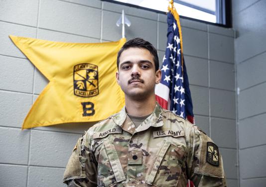 Tanis Arnold is wearing an Army uniform and standing in front of two flags