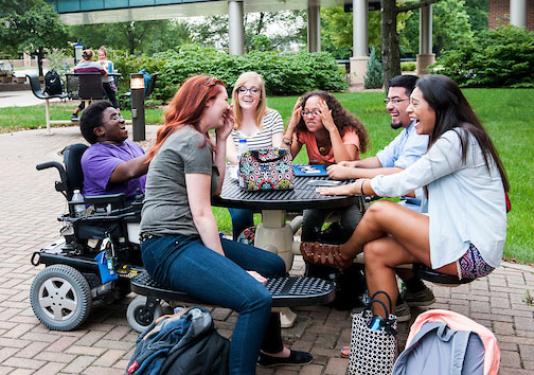 A group of laughing students sitting at a table outside the student union. One of the students is in a wheelchair.